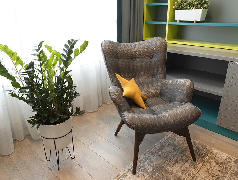 Un macetero alto con patas junto al sillón de un salón. Tiene una planta artificial de color verde.